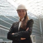Female engineer in hard hat and suit, stood with arms folded smiling down at the camera