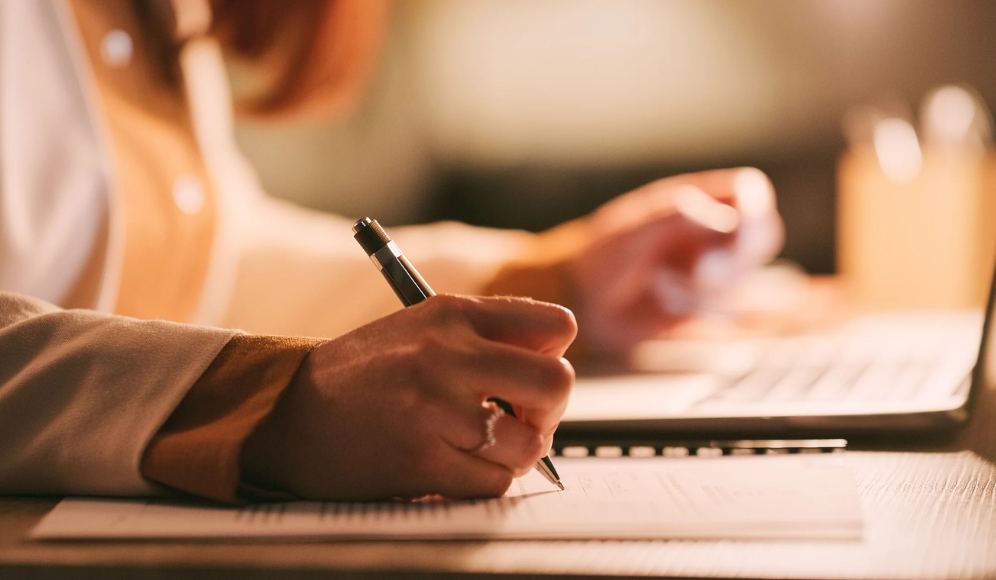 A side view of a hand writing on paper, and the other hand resting on a laptop